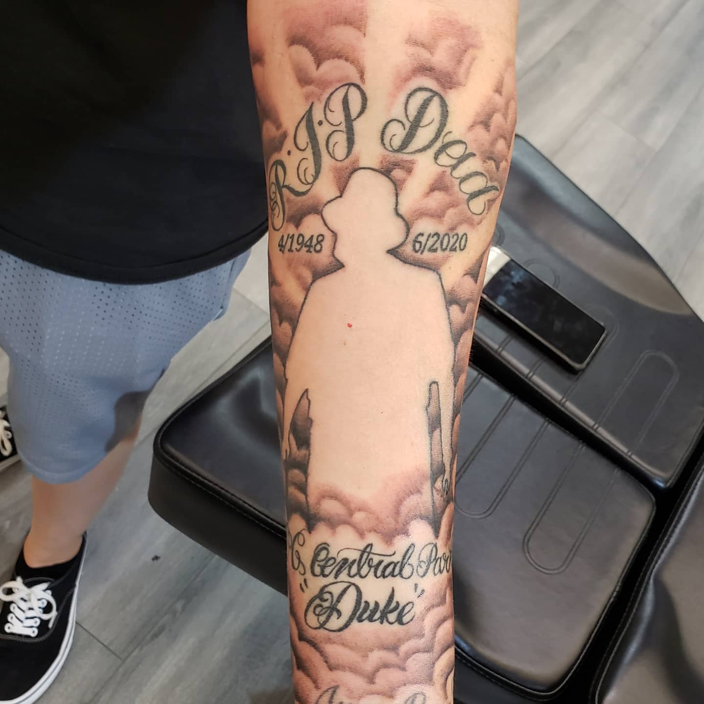 Forearm tattoo of a figure with text "RIP Dad" and dates, clouds, and "Central Park 'Duke'